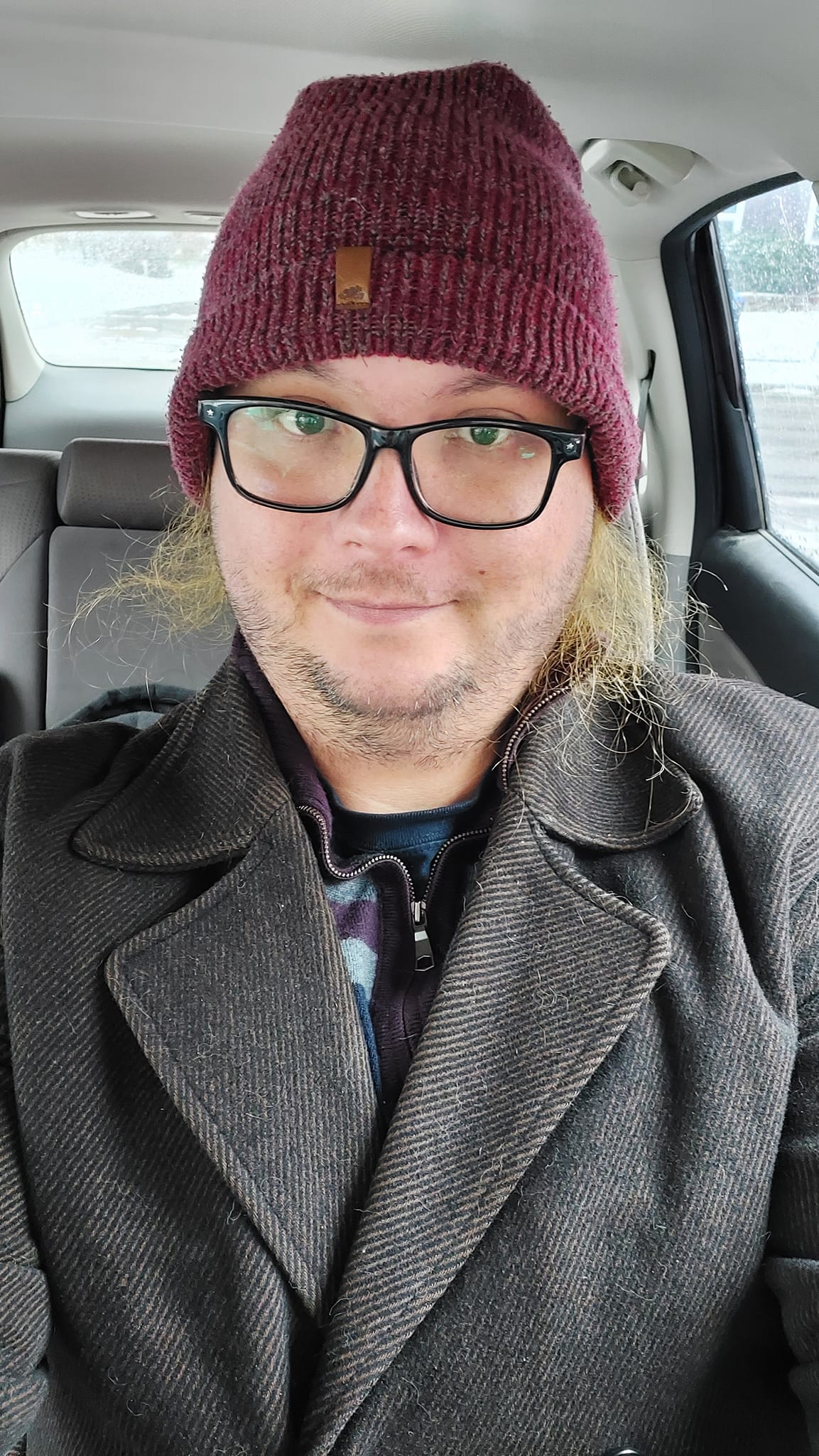 A man with green hair is wearing a black peacoat and red toque while sitting in a car, it is bright outside